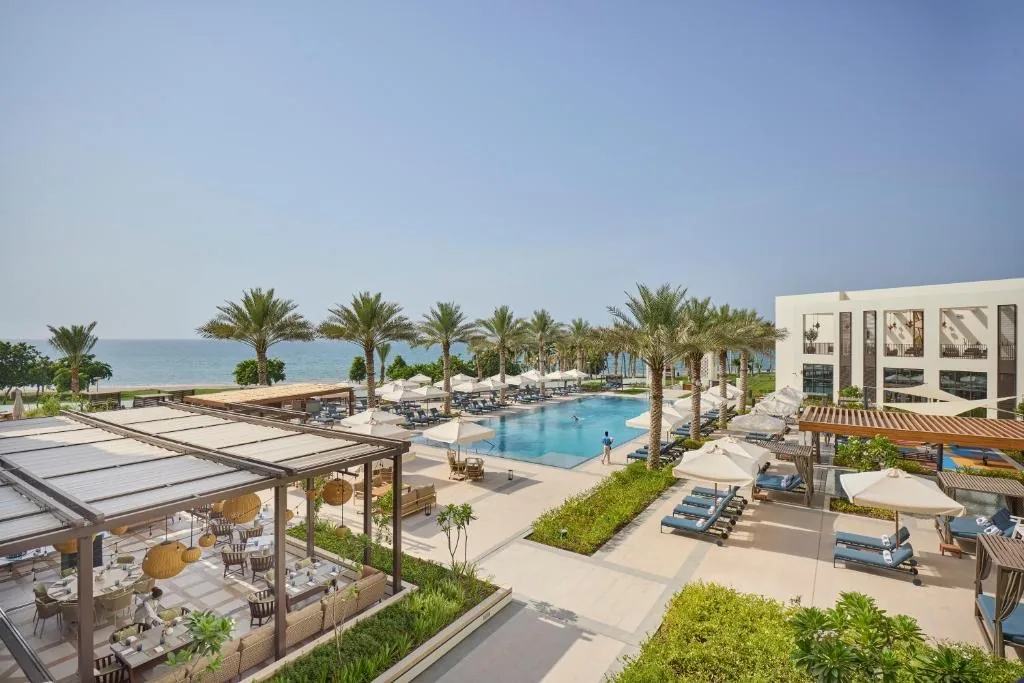Mandarin Oriental, Muscat image: a palm-lined pool terrace with sun loungers, white umbrellas and a shaded dining pavilion, the beach and the Gulf of Oman beyond, and a white residential block on the right